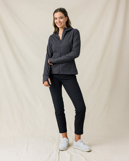 A woman modeling a dark gray zip-up athletic jacket and black cropped pants with white sneakers, standing against a draped beige fabric backdrop.