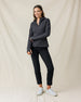 A woman modeling a dark gray zip-up athletic jacket and black cropped pants with white sneakers, standing against a draped beige fabric backdrop.