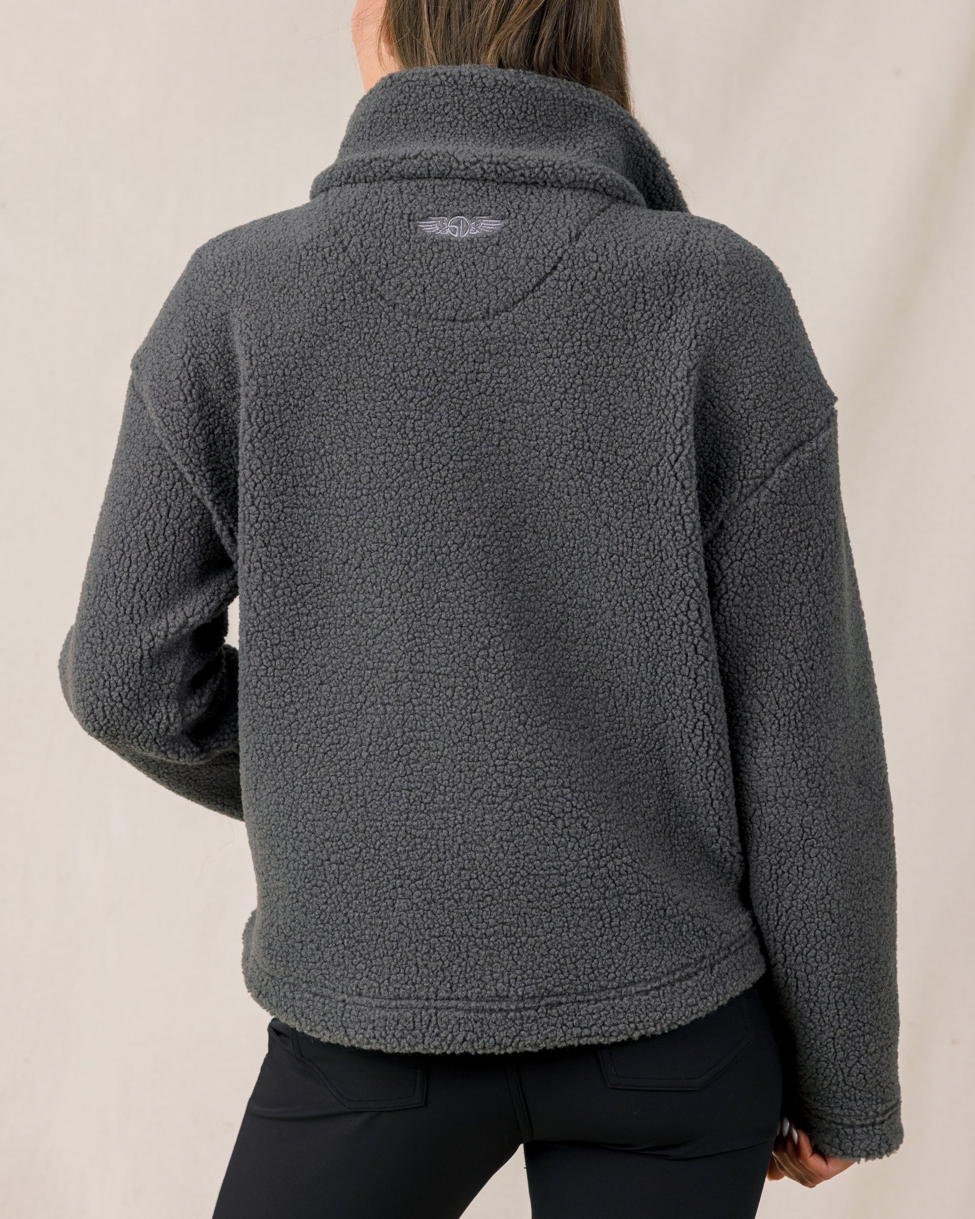The back view of a woman modeling a dark gray sherpa fleece quarter-zip jacket and black pants against a plain light beige studio background.