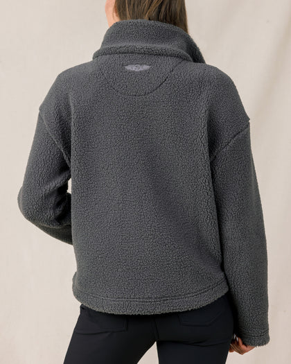 The back view of a woman modeling a dark gray sherpa fleece quarter-zip jacket and black pants against a plain light beige studio background.