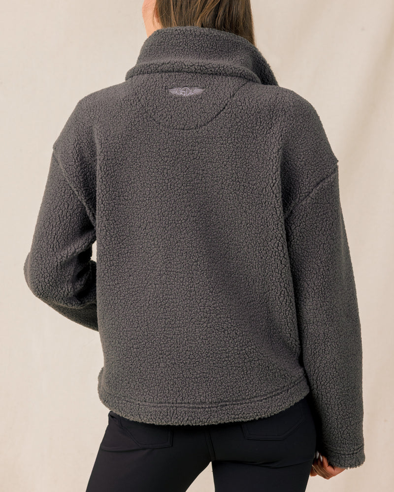 The back view of a woman modeling a dark gray sherpa fleece quarter-zip jacket and black pants against a plain light beige studio background.