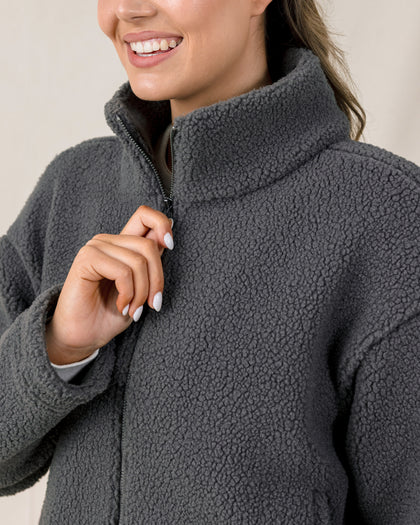 A close-up of a woman wearing a dark gray textured fleece zip-up jacket, smiling and pulling the zipper with her hand.