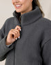 A close-up of a woman wearing a dark gray textured fleece zip-up jacket, smiling and pulling the zipper with her hand.