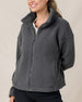 A woman wearing a dark gray sherpa fleece zip-up jacket with a high collar, paired with a white shirt and black pants, against a plain beige background.