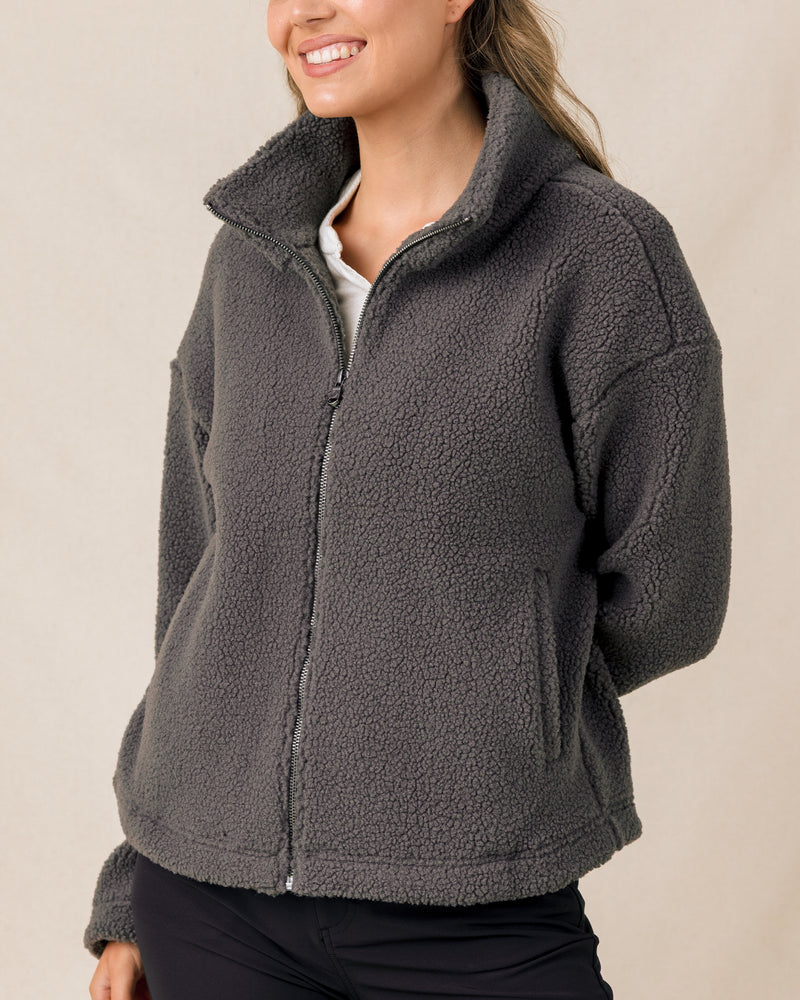 A woman wearing a dark gray sherpa fleece zip-up jacket with a high collar, paired with a white shirt and black pants, against a plain beige background.