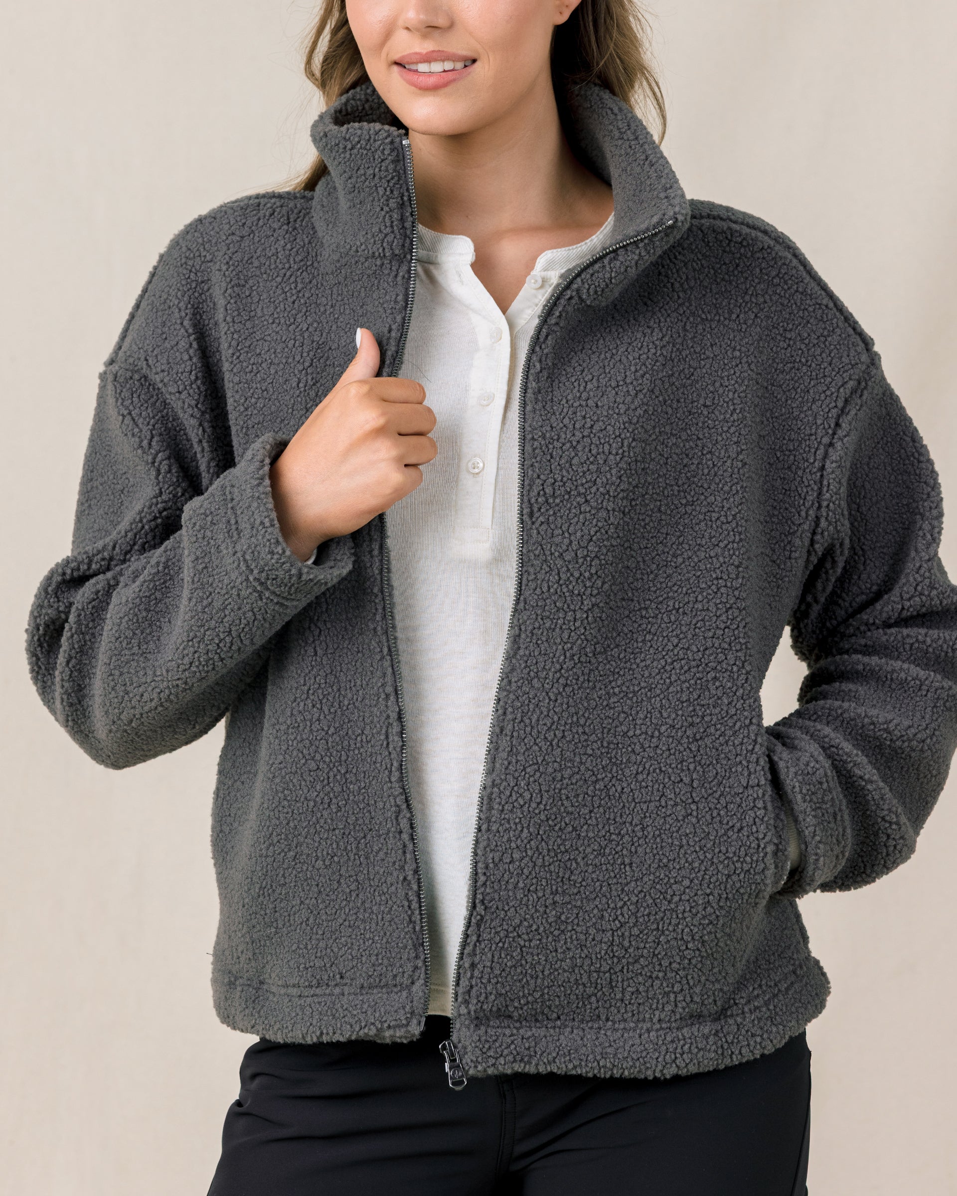 A woman modeling a dark gray fleece zip-up jacket over a white shirt and black pants, shown from the nose down against a plain beige studio background.