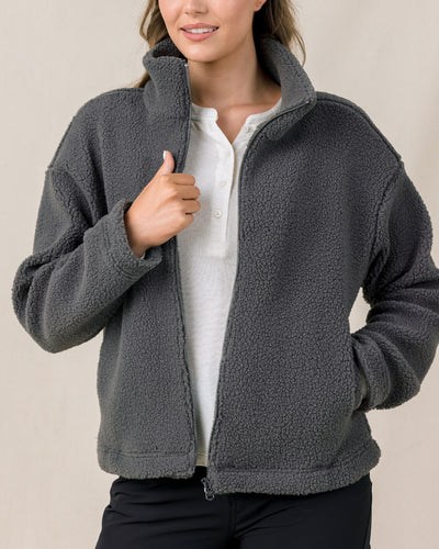 A woman modeling a dark gray fleece zip-up jacket over a white shirt and black pants, shown from the nose down against a plain beige studio background.