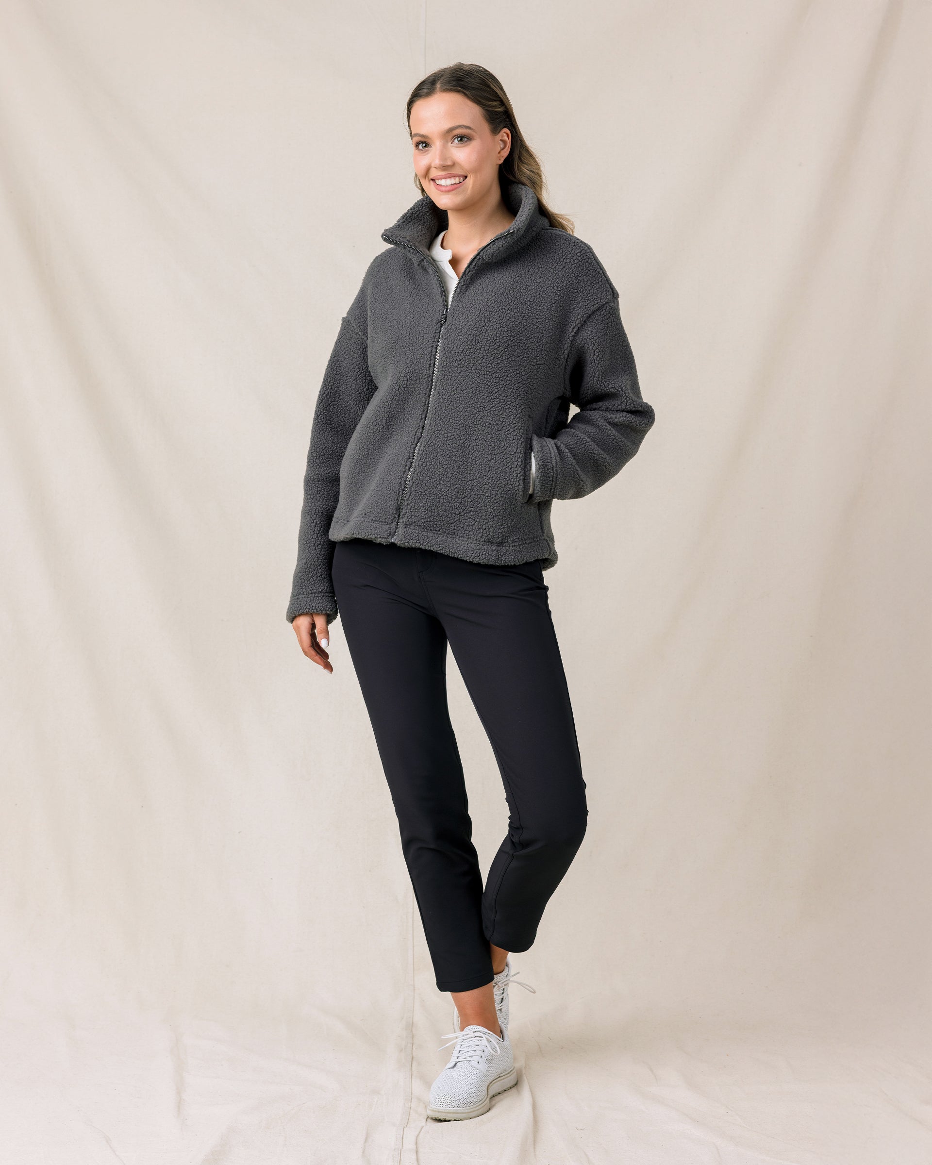 A woman modeling a dark gray fleece zip-up jacket, black cropped pants, and white sneakers against a draped beige fabric backdrop.