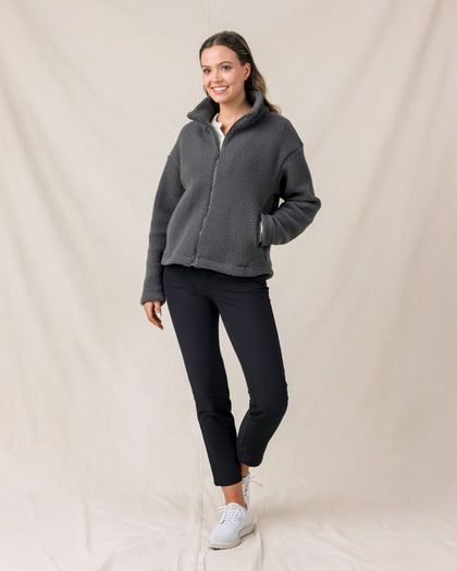 A woman modeling a dark gray fleece zip-up jacket, black cropped pants, and white sneakers against a draped beige fabric backdrop.