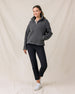 A woman modeling a dark gray fleece zip-up jacket, black cropped pants, and white sneakers against a draped beige fabric backdrop.
