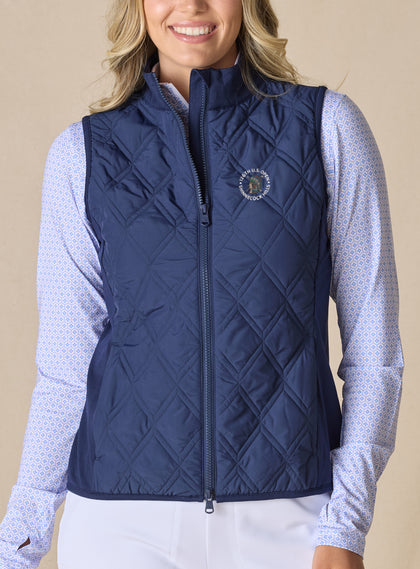 Women's 126th U.S. Open Hestia Vest in New Indigo, front model view featuring a navy quilted sleeveless vest with a full zip closure and the 126th U.S. Open Shinnecock Hills embroidered logo on the left chest layered over a white and blue printed long sleeve top with white pants