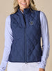 Women's 126th U.S. Open Hestia Vest in New Indigo, front model view featuring a navy quilted sleeveless vest with a full zip closure and the 126th U.S. Open Shinnecock Hills embroidered logo on the left chest layered over a white and blue printed long sleeve top with white pants