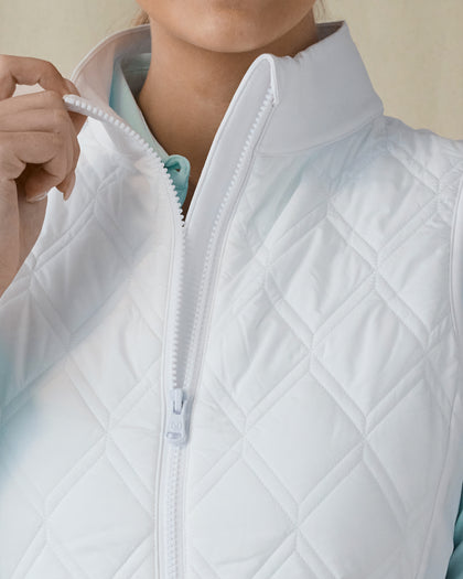 Women's Hestia Vest in White, detail close-up view featuring the diamond quilted pattern, mock neck collar, and white zipper hardware with model pulling open the zipper, layered over a mint long-sleeve top