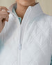Women's Hestia Vest in White, detail close-up view featuring the diamond quilted pattern, mock neck collar, and white zipper hardware with model pulling open the zipper, layered over a mint long-sleeve top