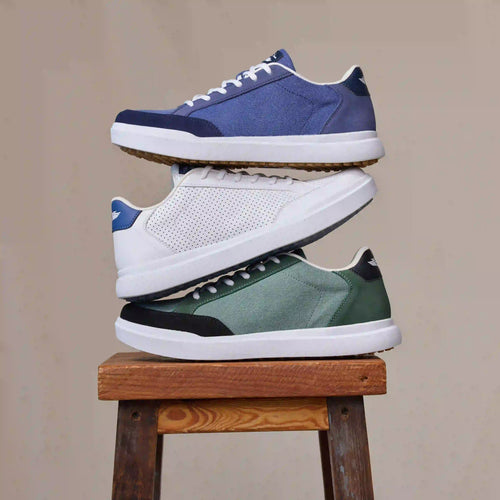 Three sneakers stacked vertically on a wooden stool against a plain beige background: a blue pair at the top, a white pair in the middle, and a green-and-black pair at the bottom.