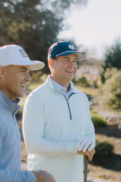 Two men smiling outdoors on a golf course. The man on the right wears a navy blue cap, a white quarter-zip pullover, and white golf gloves while holding a golf club.