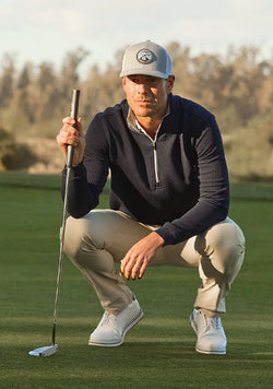 Man crouching on a golf green holding a putter, wearing a navy Optic Quarter Zip, khaki pants, white shoes, and a gray cap.
