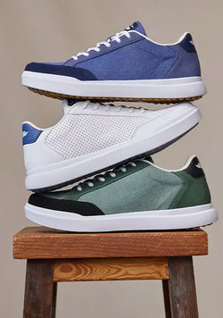 Three pairs of low-top sneakers stacked vertically on a wooden stool against a plain beige background. The top pair is blue and navy, the middle pair is white, and the bottom pair is green and black.