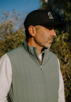 A man wearing a sage green quilted zip-up vest over a white long-sleeve shirt, paired with a black hat, photographed outdoors against a blurred background of trees and blue sky.