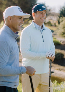 Two men smiling on a golf course, one wearing a light green quarter zip and khaki shorts with a navy cap, and the other wearing a light blue quarter zip and a white cap.