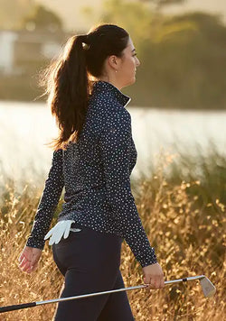 A woman in a navy long-sleeve quarter zip with white polka dots and blue golf pants walks through tall dry grass on a golf course, holding a golf club. She is seen from the back-three-quarter view in warm golden hour light.