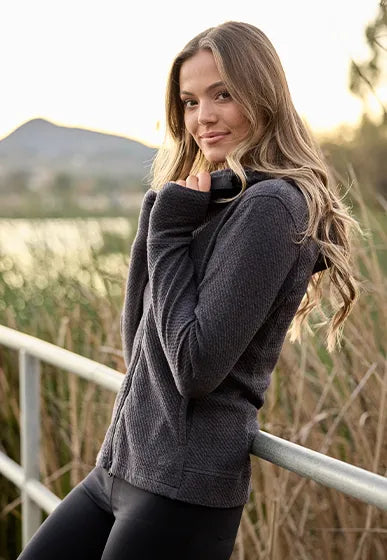 A woman wearing a dark gray textured zip-up hoodie and black leggings, leaning against a metal railing outdoors near a lake with a mountain in the background.