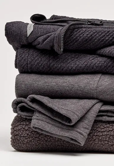 A stack of folded dark gray fleece and knit garments, including a zippered jacket and soft sweaters, photographed on a clean light gray studio background.