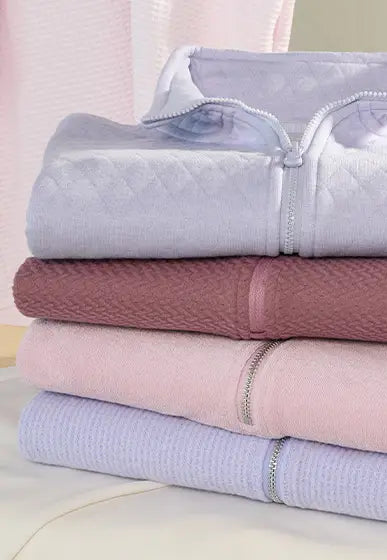 A stack of four folded quilted zippered blankets or towels in pastel shades of lavender, dusty rose, light pink, and pale blue, arranged on a light surface.